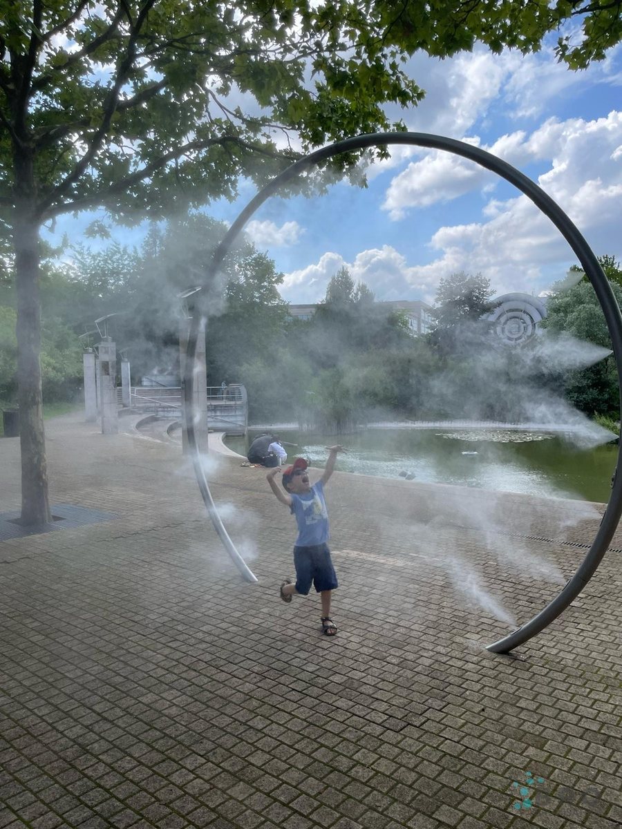 Enfant les bras levés sous une arche de brumisation BRO, au bord d'un bassin, un jour d'été