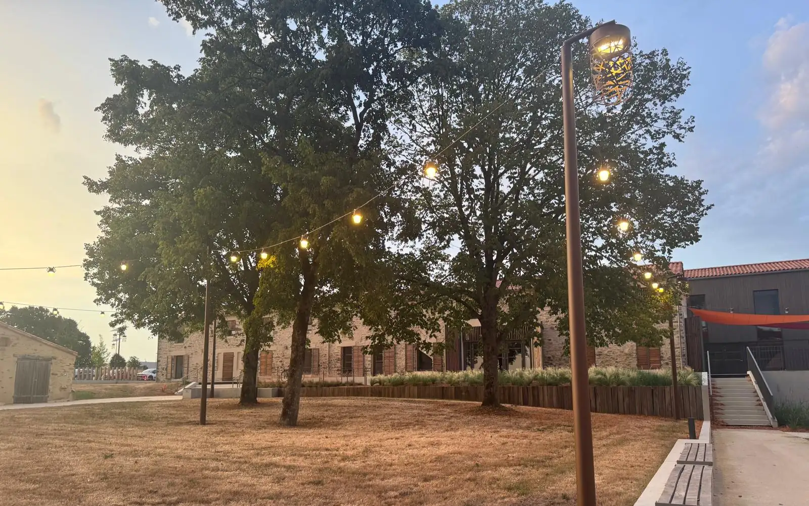 Place arborée du Plessis-Richard éclairée par une lanterne sur mât et une guirlande entre les arbres, à la tombée du jour
