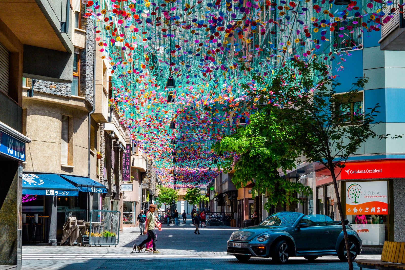Rue commerçante entière couverte d'un plafond climatique de fleurs colorées, passants et terrasses à l'ombre en plein jour